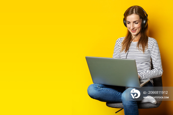 Young woman in headphones sitting on chair and holding laptop on isolated yellow background