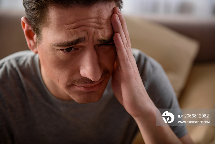 How it hurts. Close up portrait of exhausted man suffering from headache. He is upset and sitting in bedroom. Focus on his face