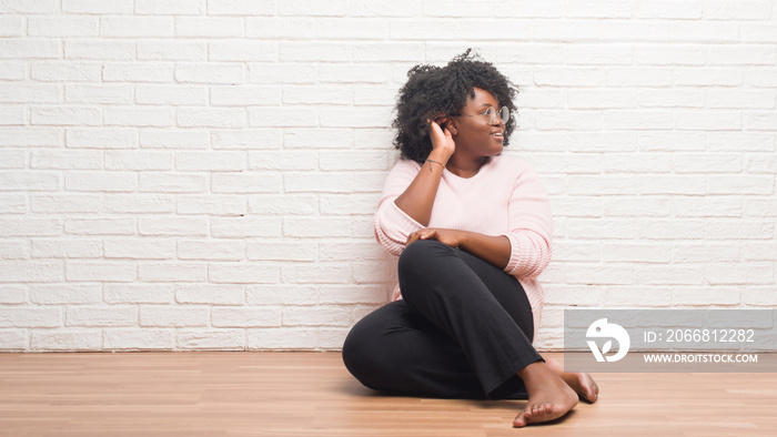 Young african american woman sitting on the floor at home smiling with hand over ear listening an hearing to rumor or gossip. Deafness concept.