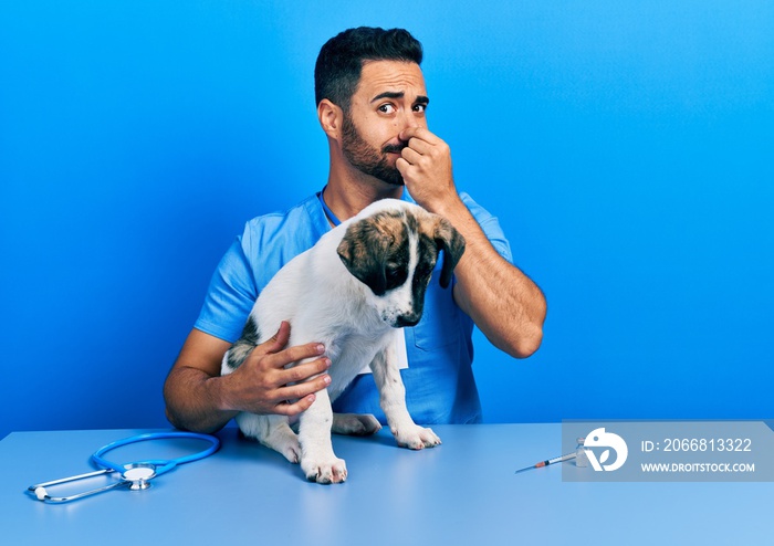 Handsome hispanic veterinary man with beard checking dog health smelling something stinky and disgusting, intolerable smell, holding breath with fingers on nose. bad smell