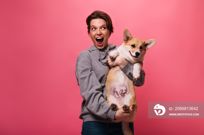 Joyful woman in grey hoodie holding corgi on pink background. Emotional teen girl in jeans plays with dog on isolated