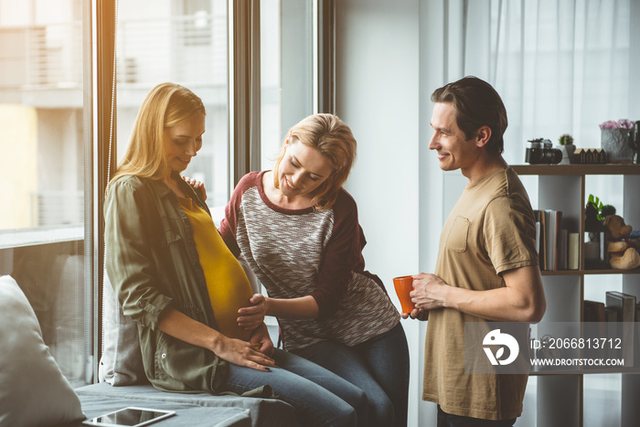 Soon you will be with us. Happy woman is talking with unborn baby and smiling. She is touching belly of surrogate expectant mother with love while man is standing near them