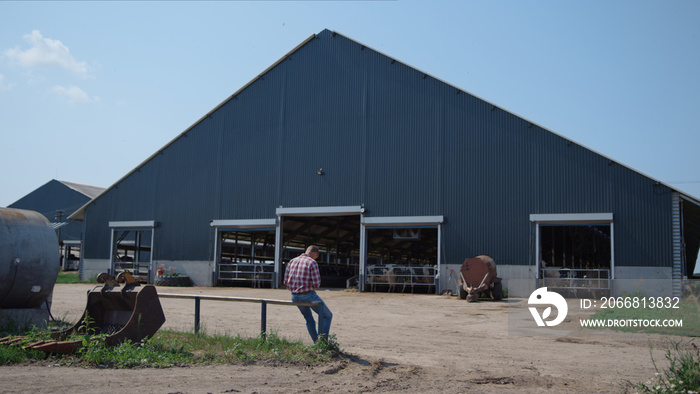 Ranch worker sitting outside big cowshed on fence after feeding livestock herd.