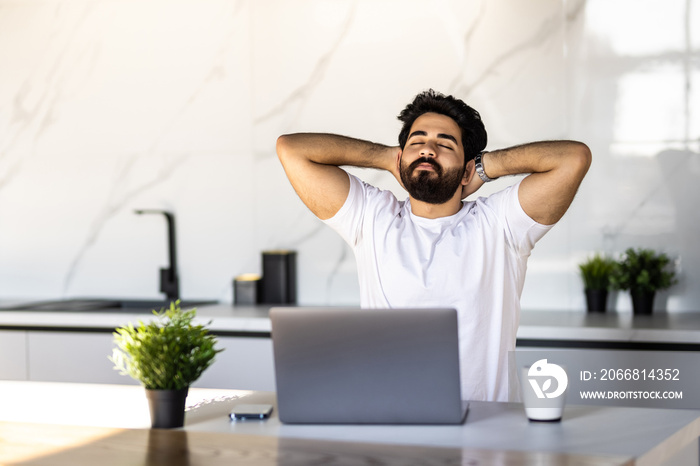 Relaxed freelancer man leaning back in chair, resting after work with laptop and holding hands behind head, sitting in kitchen interior, working distantly from home