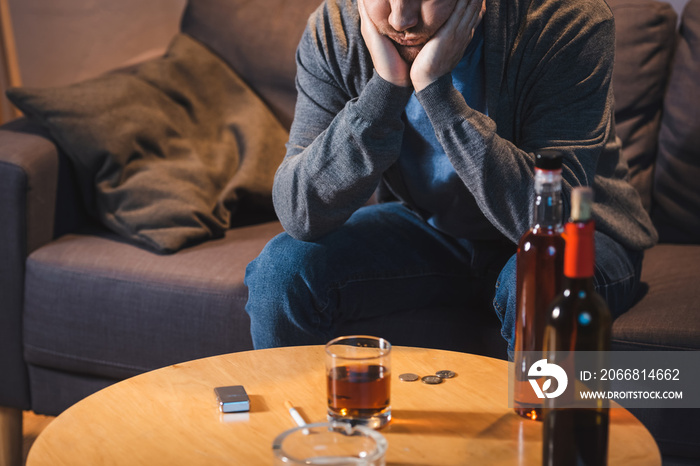 Drunk man sitting near table with glass and bottles of alcohol at home