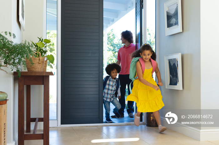 Multiracial young man standing at entrance while children with backpacks running inside house