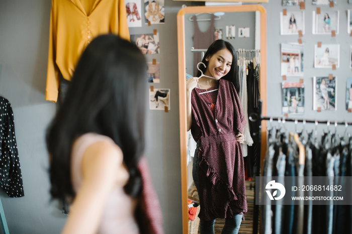 shopping, fashion, style and people concept - happy woman choosing clothes and looking to mirror in mall or clothing store