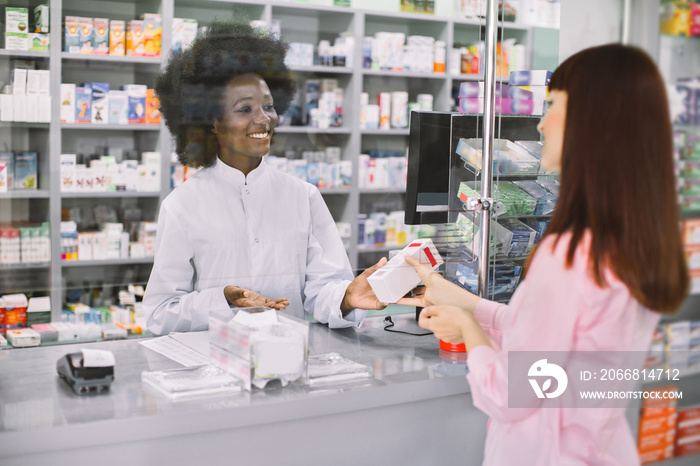 Happy young African female pharmacist standing behind the counter and offering medicine box, talking with young woman customer. Medicine sale, healthcare concept