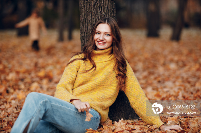 Young woman model sitting in autumn park with yellow foliage maple leaves. Fall season fashion