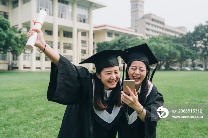 Joy girl friends using speaker option on smart phone. cheerful ladies graduate students standing outdoor having fun laughing on lawn green grass summer talking on cellphone holding diploma degree.