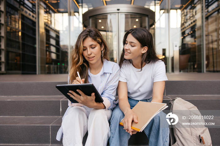 Calm young caucasian girls use tablet to search for information sitting on street. Blonde and brunette are preparing for lesson, wear casual clothes. Lifestyle concept