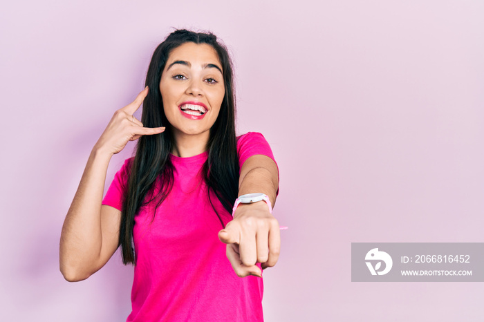 Young hispanic girl wearing casual pink t shirt smiling doing talking on the telephone gesture and pointing to you. call me.