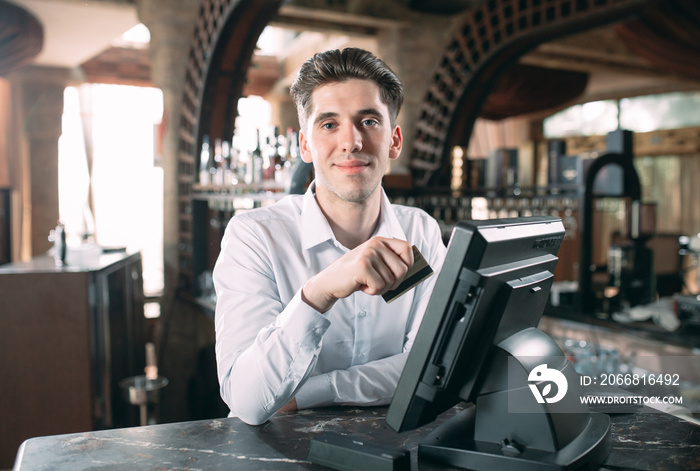 small business, people and service concept - happy man or waiter in apron at counter with cashbox working at bar or coffee shop.