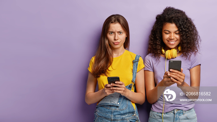 Dissatisfied young woman has no internet connection on mobile phone, cheerful African American girl has amusing chat in network, listens music in headphones isolated on purple wall. Technology concept