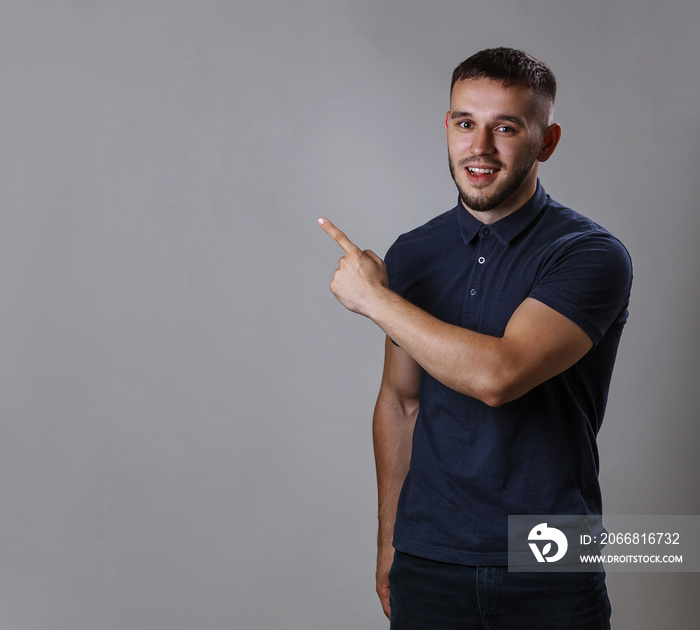 Portrait of funny man wearing a shirt pointing finger to side show or demonstrating something while standing on gray background copy space