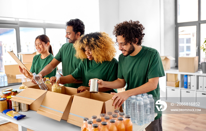 charity, donation and volunteering concept - international group of happy smiling volunteers packing food in boxes according to list on clipboard at distribution or refugee assistance center