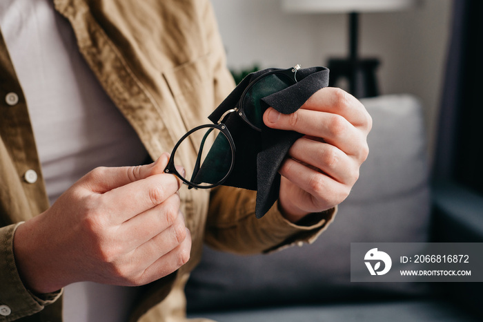 Close up of male hands cleaning eyeglasses with small black cloth sitting alone on cozy couch in living room at home. Cleaning glasses concept