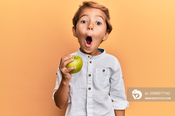 Adorable latin kid holding green apple scared and amazed with open mouth for surprise, disbelief face