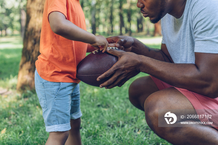 cropped view of african american father showing rugby ball to son in park