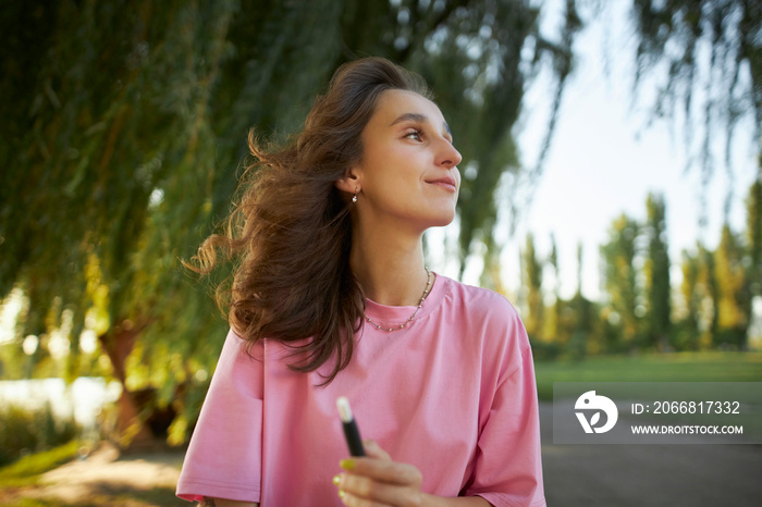 Cheerful girl in a pink t-shirt with an electronic cigarette in the park. Beautiful girl in a pink t-shirt, beaming smile, holding an electronic cigarette. Teenage girl using an electronic cigarette.