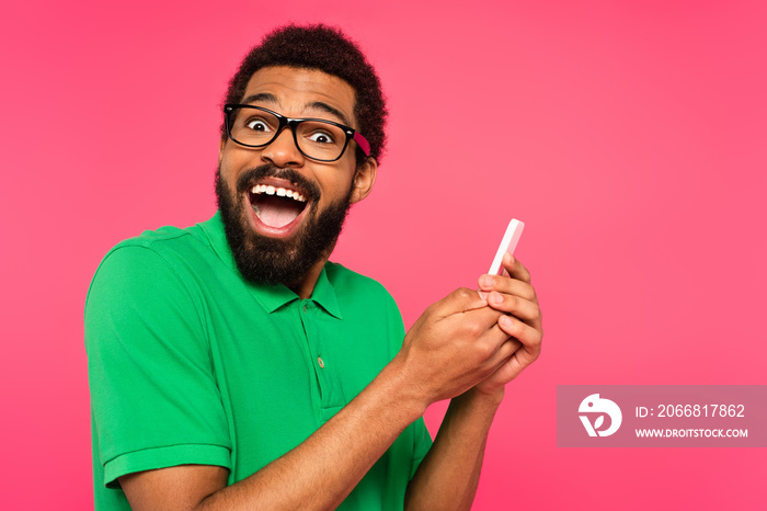 amazed african american man in green t-shirt using smartphone isolated on pink