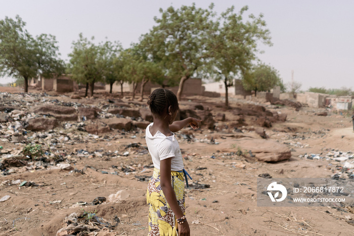 Young african girl standing with her back to the camera pointing to the desolate landscape of her homeland characterized by climte change, desertification and waste pollution