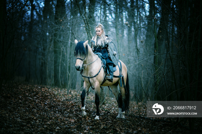 Warrior Woman on a horse in the woods. Scandinavian viking riding horse with ax in hand, blonde hair in a traditional clothes with fur collar, war makeup, forest.