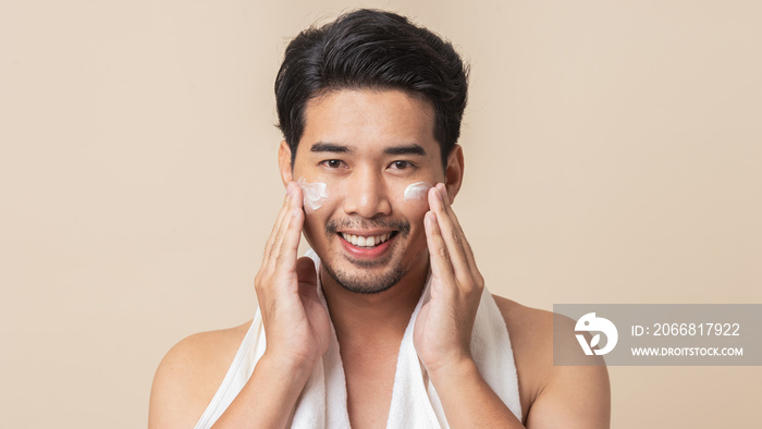 Young Asian man happy smiling and applying moisturizer skin care cream on face after take a shower on brown background. Asian man with mustaches. Men’s skin care.