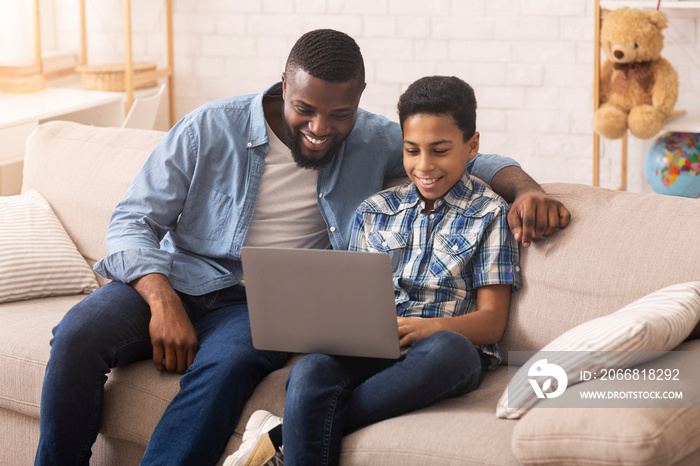 Father Teaching Son To Use Laptop, Sitting On Couch At Home