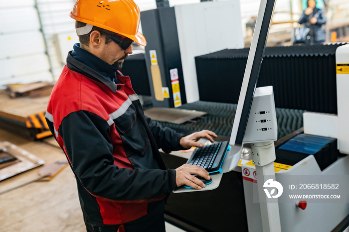 A worker in a hard hat and overalls operates a laser in a production shop. Rolled metal