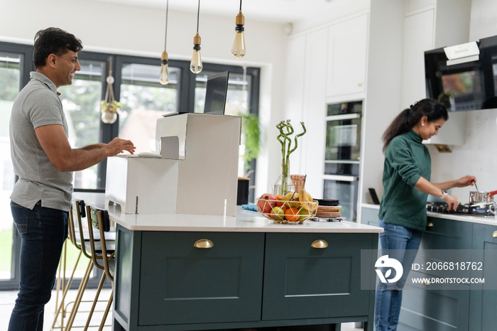 Man working on laptop in kitchen while wife preparing meal