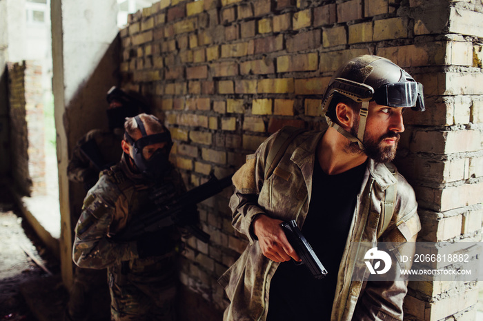 A picture of serious bearded man hiding behind wall and looking to the side. He has gun. Other two men in maska are standing behind him. They are waiting for his command to start a fight.