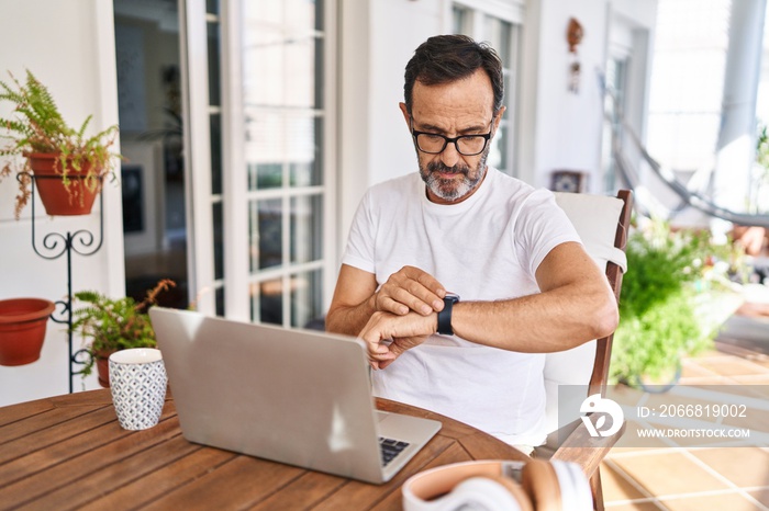 Middle age man using computer laptop at home checking the time on wrist watch, relaxed and confident