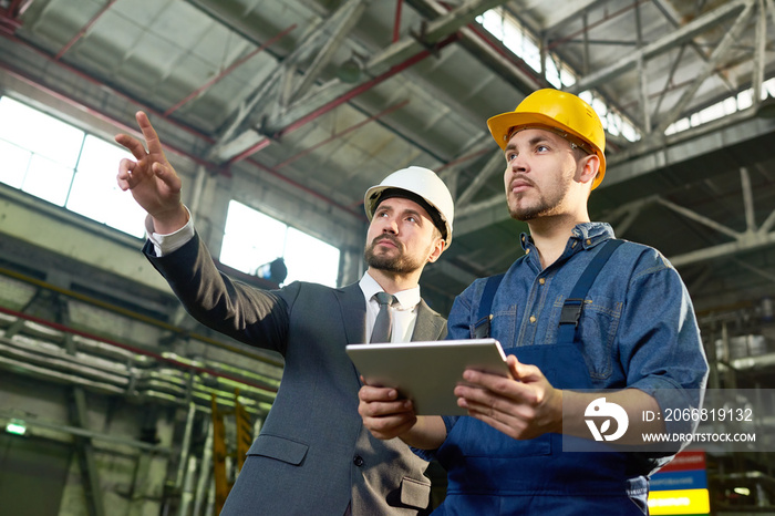 Waist up portrait of handsome businessman pointing up while discussing something with factory worker holding digital tablet, copy space