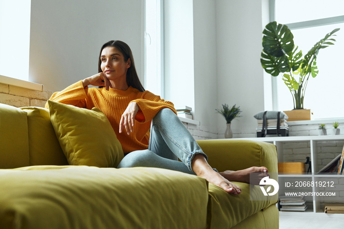 Thoughtful young woman holding hand on chin while sitting on the couch at home