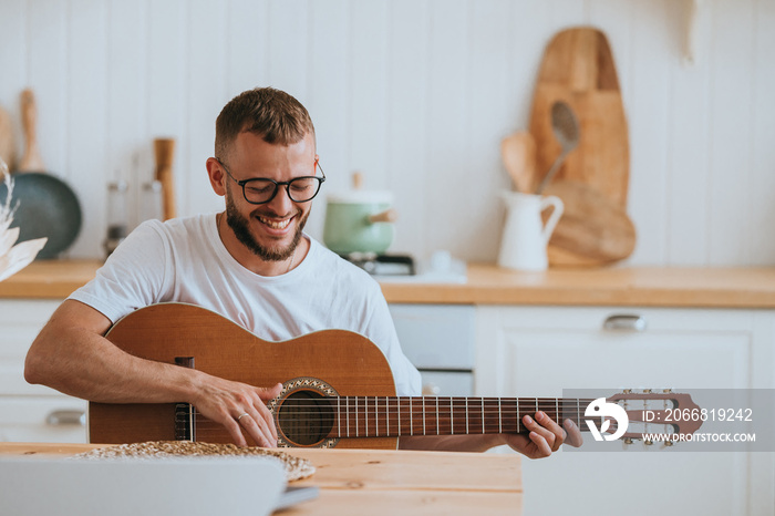 Cheerful Australian beardy guy sitting at kitchen plays guitar toothy smiles enjoys music, singing song on vacations. Handsome caucasian young man practicing at home. Hobbie, musician.