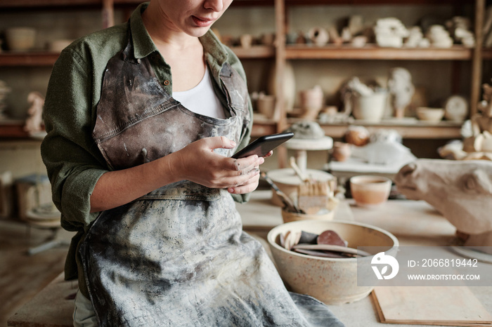 Close-up of woman in apron working online on her mobile phone while working with clay in the workshop