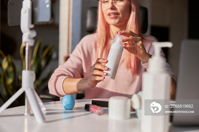 Pretty woman beauty blogger with pink hair shows bottle of cosmetic product at table