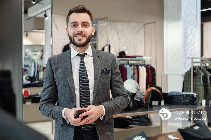 Portrait of positive handsome young businessman with beard holding smartphone in clothing shop