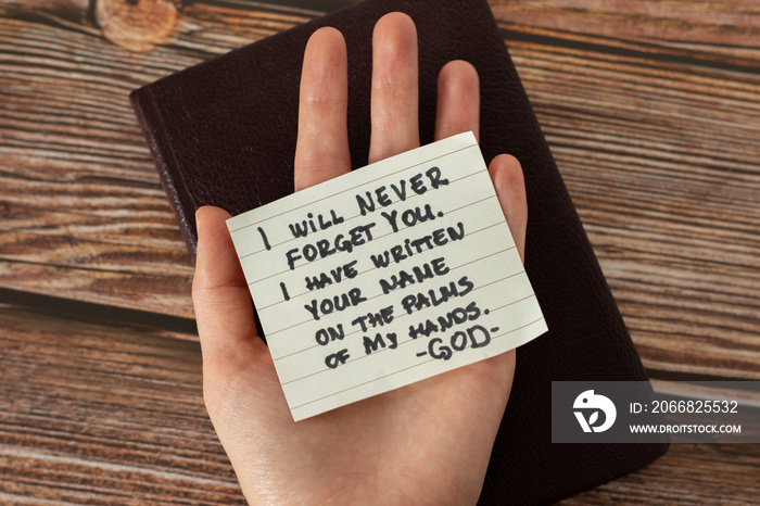 Human hand holding handwritten Bible quote about God Jesus Christ’s love and faithfulness on a wooden background. Top view. A closeup.