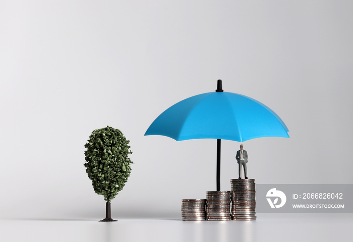 Miniature businessman standing on a pile of coins with umbrella next to a miniature tree.