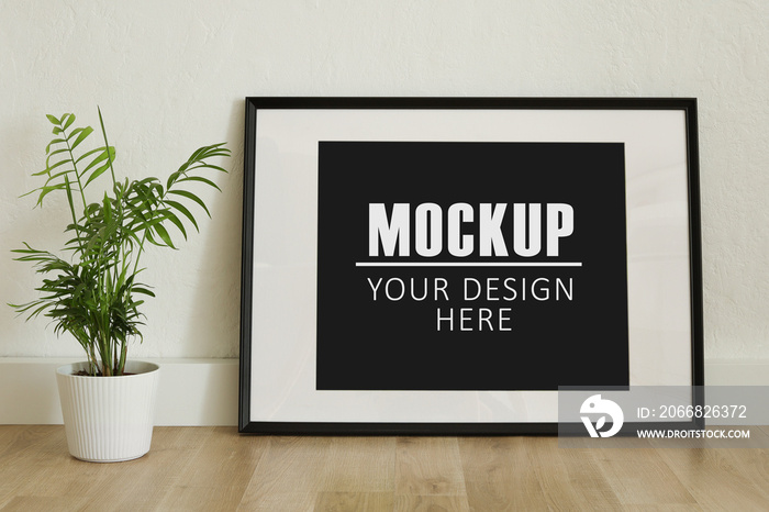 Frame, poster mock up with horizontal black frame. Empty frame standing on the wooden floor with a green plant. Free space for your picture or text, copy space. Minimalist design.