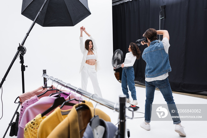 trendy model posing with raised hands near photographer, assistant with electric fan and rack with clothes.