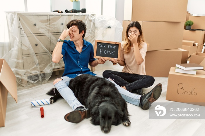 Young caucasian couple with dog holding our first home blackboard at new house smelling something stinky and disgusting, intolerable smell, holding breath with fingers on nose. bad smell