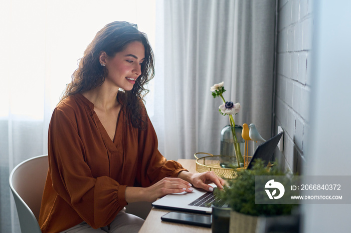 Pretty girl in brown blouse looking at laptop screen during network in home office while sitting against window with grey curtains