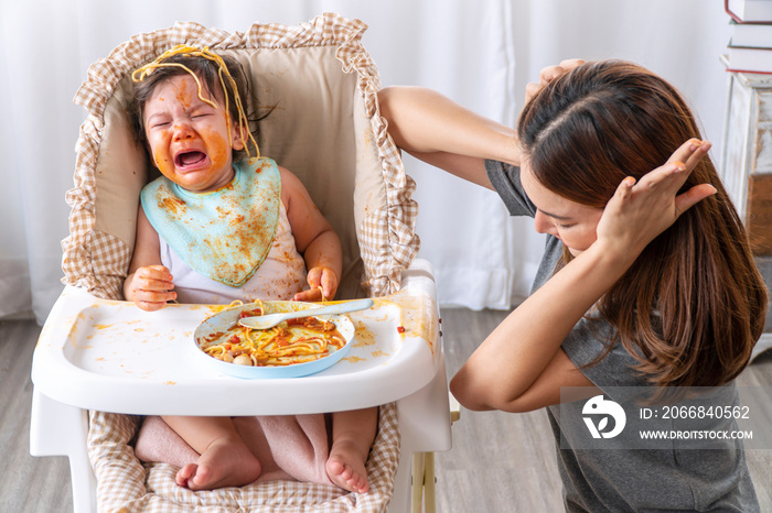 Unhappy toddler child with tomato sauce on her face crying with her mother sitting together beside in the room. Upset messy little girl mix race hungry eating spaghetti with mom at home.