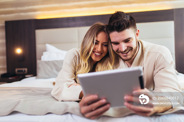 Happy couple using digital tablet while lying together in bed in hotel room.