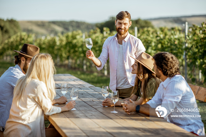 Group of a young people drinking wine and talking together while sitting at the dining table outdoors on the vineyard on a sunny evening