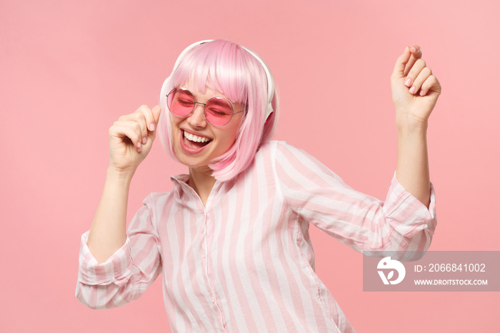 Young happy laughing girl with pink hair and glasses, listening to music in wireless headphones and dancing, isolated on studio background