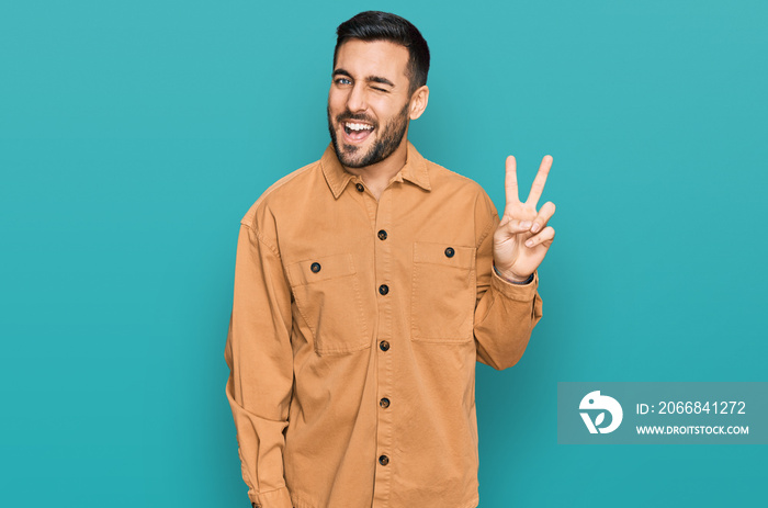 Young hispanic man wearing casual clothes smiling with happy face winking at the camera doing victory sign. number two.